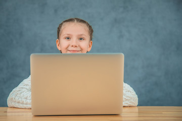 Little cute blonde girl with brainds is smiling and sitting in front of grey laptop.