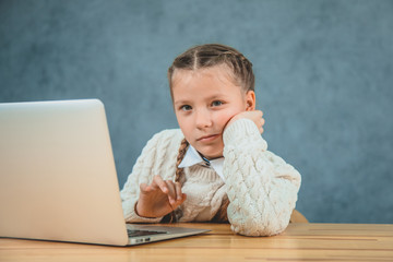 Cute schoolgirl with blond hair and braids is looking seriously near grey laptop.