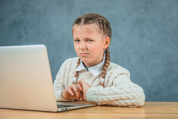 Beautiful, charming schoolgirl with blond braids is sitting in front of the laptop and makes faces.