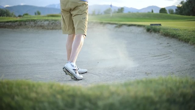 Slow motion shot behind a golfer hitting the golf ball out of the sand trap onto the putting green. The golfer is playing on a nice resort golf course.