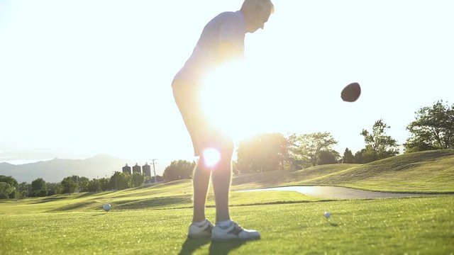 Slow motion shot of a male on a resort golf course about to drive a golf ball off of the teebox. Cinematic sun flares are happening behind the man.