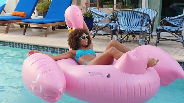 At the swimming pool, a beautiful woman on top of a giant flamingo floatie pushes herself off the edge to drift off with a smile.