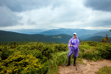 Naklejka premium The girl, a tourist with Scandinavian sticks in a raincoat on the background of the beautiful Carpathian mountains. Rise in the mountains. Travel concept, leisure activity, vacation