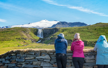 Fototapeta na wymiar Three friends wearing shirts, colorful jackets, have relationship and trekking to see waterfall views traveling together, watching a mountain view and feel freedom summer in Iceland.
