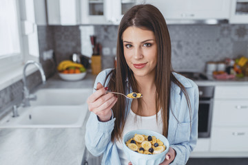 Attractive brunette woman in blue shirt eating bowl of cereal and smiling at the camera in the kitchen at home.