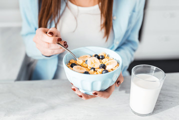 Detail of a young woman sitting on a living room couch, holding a bowl of cereal and having breakfast