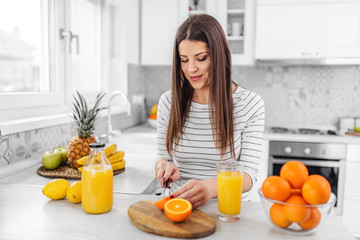 Smiling young woman in a gray T-shirt squeezes a fresh orange on fruit juice. Healthy eating concept. In the interior of the kitchen