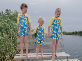 Fototapeta premium Funny kids in bathing suits sit together on wooden bridge near river. boys are happy on vacation in village together. Summer day, river, swimming in the water