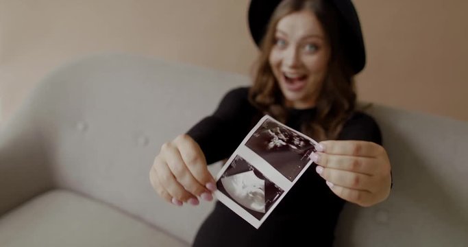 Beautiful caucasian pregnant young woman sits on studio sofa, shaking ultrasound images and smiling happy emotions. 4K RAW graded footage 24fps