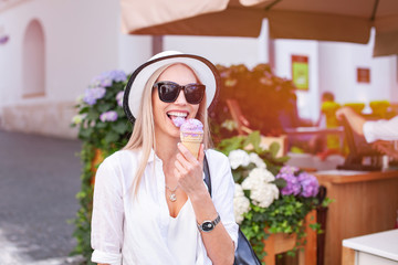 Beautiful young woman in hat and glasses walking on the streets of a European city. Lovely girl traveling in Europe. Perfect blonde European woman eating blueberry icecream cones and enjoy life.