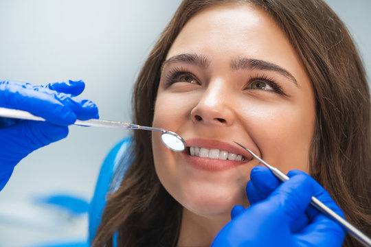 smiling happy brunette woman patient examined by dentist in blue gloves using dental mirror and scaler sitting in dental clinic