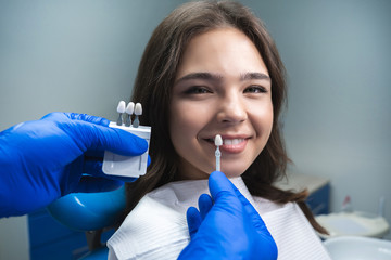 dentist in blue medical gloves applying sample from tooth enamel scale to smiling woman patient to pick up right shade for teeth bleaching procedure