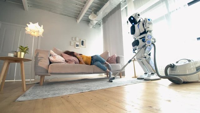Human-like robot is cleaning the room with a woman sitting on a sofa