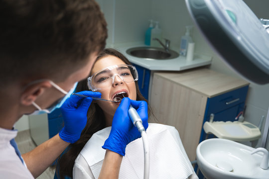 dentist in mask filling the patient's root canal while she is lying on dental chair wearing safety glasses under the medical lamp in clinic