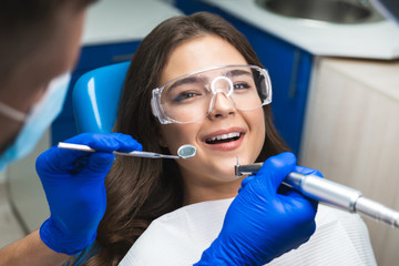 smiling happy brunette woman patient examined by dentist in blue gloves and medical mask using dental mirror and scaler sitting in dental clinic