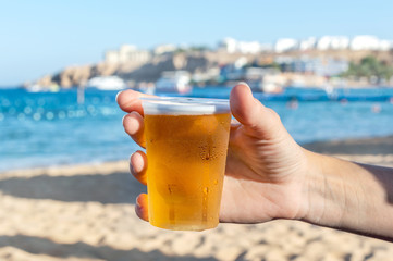 Woman's hand holding plastic glass of beer on sea beach.