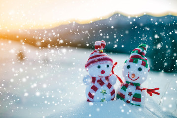 Two small funny toys baby snowman in knitted hats and scarves in deep snow outdoors on blurred mountains landscape background. Happy New Year and Merry Christmas greeting card theme.