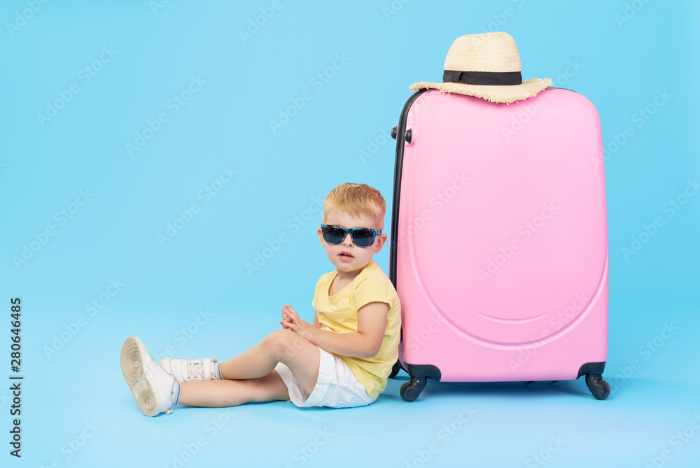 Wall mural Happy child boy tourist packs clothes into a suitcase for travel, summer vacation. Toddler kid in sunglasses sits near a pink suitcase on a blue background