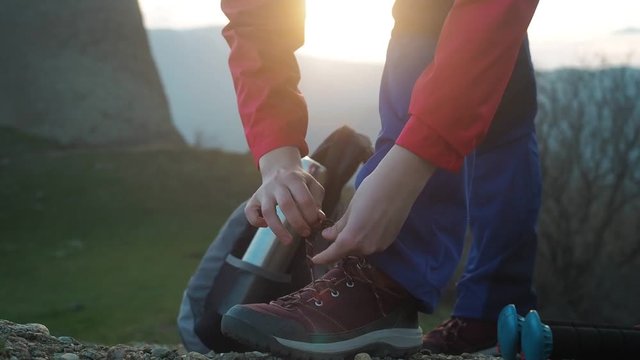 Young wiman hiker tying shoelaces, beautiful sunset in mountains on the background. Female hiking boots close up, feet walking, trekking national park trail.