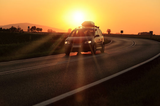 Samoch&oacute;d z bagażnikiem dachowym na zakręcie drogi o zachodzie słońca.