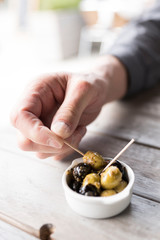 hand is grabbing, black and green olives in white bowl with wooden stick.