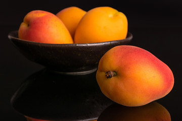 Group of four whole fresh orange apricot , one is in the front and the other three are in the back in a dark ceramic bowl isolated on black glass