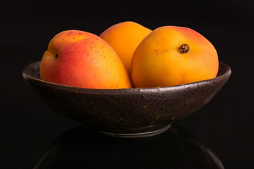 Group of three whole fresh orange apricot in a dark ceramic bowl isolated on black glass