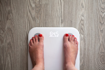 Woman stands on white modern electronic scales, which show 64.5 kg. Only feet are visible. Scales stand on gray wooden floor. Space for text. Healthy lifestyle, diet, weight loss concept. Horizontal.