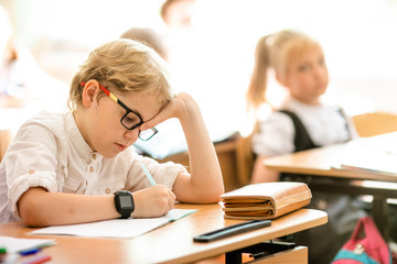 Blonde boy with big black glasses sitting in classroom, studing, smiling. Education on elementary...