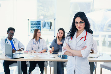 Smiling medical doctor woman with stethoscope standing in front of medic team at hospital. Attractive young female doctor in white coat and glasses looking at the camera.