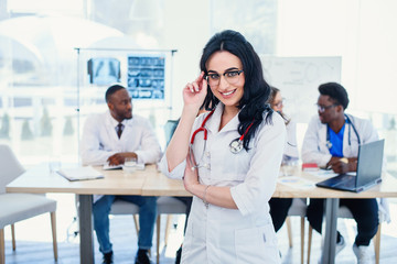 Fototapeta premium Smiling medical doctor woman with stethoscope standing in front of medic team at hospital. Attractive young female doctor in white coat and glasses looking at the camera.