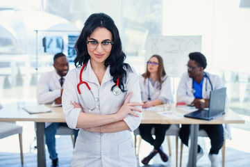 Fototapeta premium Smiling medical doctor woman with stethoscope standing in front of medic team at hospital. Attractive young female doctor in white coat and glasses looking at the camera.