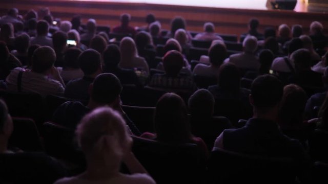 Full auditorium of people. Spectators watch performance show or view in theater, back view. Staging in theatre. Many people in playhouse sitting in chairs and looking at scene.
