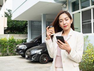 Fototapeta premium Asian woman holding coffee cup and using smartphone while standing in front of house. Portrait of smiling asian businesswoman