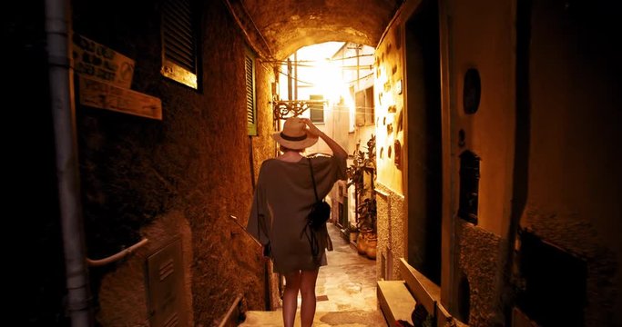 Romantic young woman in the wonderful narrow street admiring sunset. Back view of pretty girl wanderer walking through Rio Majore village on summer tour. Italian destination.