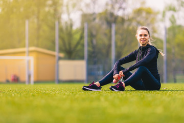 Fototapeta premium young pretty thirsty blonde Caucasian girl in suit resting after sport run in the park on football stadium on grass drinking water and smiling enjoying and listening music with her wireless headphones
