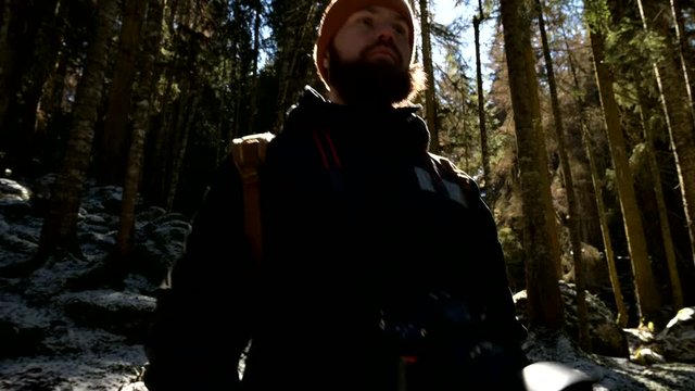 Portrait of a serious bearded man photographer with a backpack and a camera standing in a coniferous winter forest in backlight and sunlight