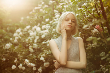 Portrait of beautiful young girl with hands on her face in white roses. Blond platinum girl with fair complexion. Sunset backlight. Image with selective focus and toning