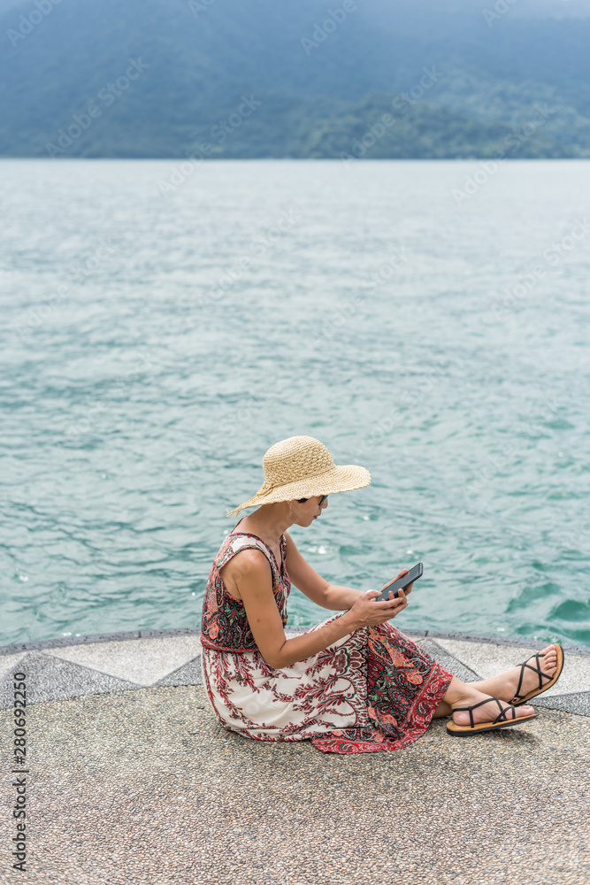 Poster woman with hat sit at a pier using cellphone