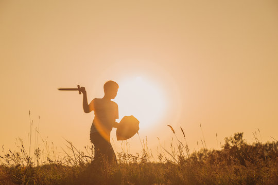 Black silhouette of cute young caucasian kid isolated on sunny orange sunset or sunrise sky background playing sword with invisible enemy. Horizontal color photography.