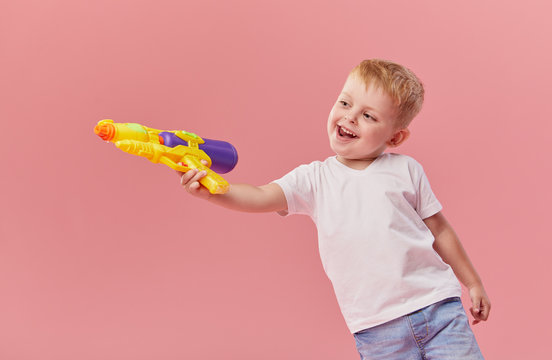 Happy little toddler boy  in summer clothes holds toy water gun isolated on pink wall background. Children studio portrait. People childhood lifestyle concept.