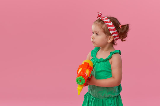Happy little toddler  girl  in summer clothes holds toy water gun isolated on pink wall background. Children studio portrait. People childhood lifestyle concept. 