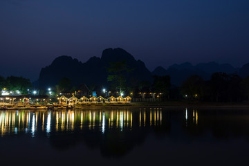 Few people sitting at a lit waterfront restaurant by the the Nam Song River and silhouette of karst limestone mountains in Vang Vieng, Vientiane Province, Laos, at dusk. Copy space.