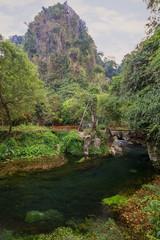 Fototapeta premium Beautiful landscape of a hill, small river and lush vegetation next to the Tham Chang (or Jang or Jung) Cave in Vang Vieng, Laos.