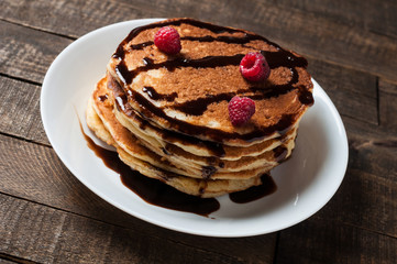 Heap of homemade pancakes with chocolate cream, raspberry lies on round white ceramic plate and old rustic wooden table