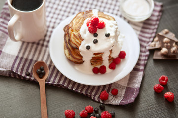 Pancackes with sour cream and tea on dark surface, top view of table decorated with muffins, raspberries, blueberries, spoon, chocolate, cup of coffe or tea, cotton towel. Tasty breakfast in morning.