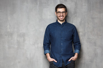 Young cheerful man wearing trendy denim casual shirt, standing against gray wall with copy space on left side
