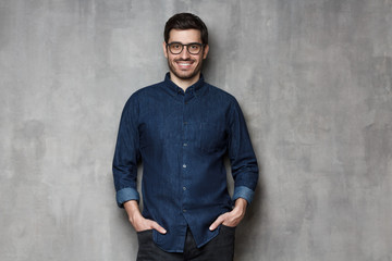 Young handsome man wearing trendy glasses and denim shirt standing leaning on gray textured wall