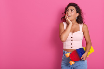 Half length shot of happy surprised curly woman, spending time with friends, ready for riding skateboard together, wears rosy t shirt and jean, posing with opene eyes. Copy space for advertisment.