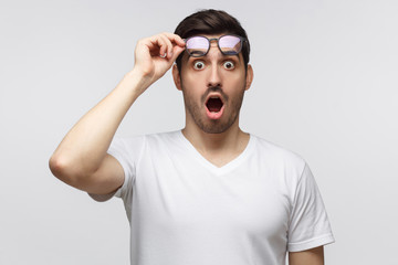 Young man in white t-shirt shouting, crabbed with shock content, holding eyeglasses with one hand